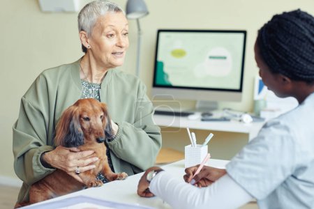 Photo for Portrait of senior woman with dog visiting vet clinic and talking to veterinarian explaining symptoms - Royalty Free Image