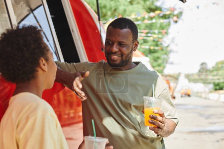 Téléchargez les photos : Le jeune père africain parle à son fils pendant qu'ils se reposent et se rafraîchissent avec des boissons fraîches après leur promenade dans le parc - en image libre de droit
