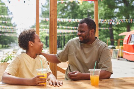 Téléchargez les photos : Papa heureux parlant et riant avec son fils pendant qu'ils buvaient du jus froid à table dans un café extérieur - en image libre de droit