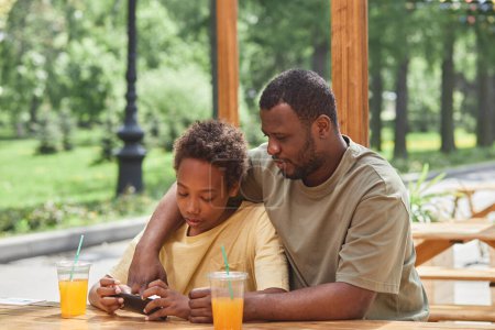 Foto de Africano niño jugando videojuego en su teléfono móvil junto con su padre mientras se sientan en la cafetería al aire libre con bebidas - Imagen libre de derechos