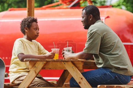 Téléchargez les photos : Papa parlant à son fils assis à la table pendant leur repos au café extérieur avec du jus froid - en image libre de droit