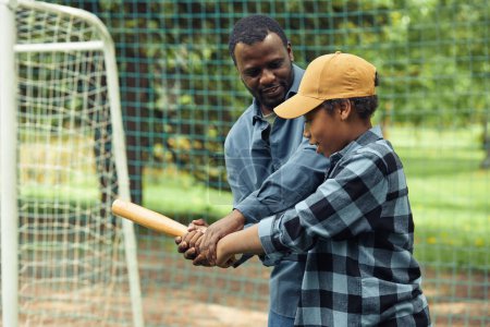 Téléchargez les photos : Père africain enseignant à son fils à tenir la batte de la bonne manière pendant leur match de baseball en plein air - en image libre de droit