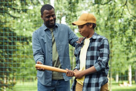 Téléchargez les photos : Africain jeune papa donnant la batte à son fils et montrant comment le tenir tout en lui apprenant à jouer au baseball - en image libre de droit
