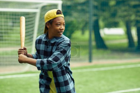 Téléchargez les photos : Garçon africain en casquette debout avec chauve-souris et prêt à frapper la balle pendant le match de baseball en plein air - en image libre de droit