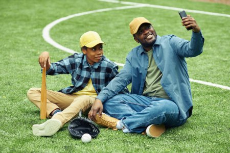 Foto de Feliz papá haciendo retrato selfie con su hijo en su teléfono móvil mientras descansan en la hierba después del partido de béisbol - Imagen libre de derechos