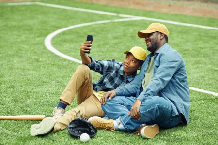 Téléchargez les photos : Africain heureux fils faire selfie portrait sur son smartphone avec son père alors qu'ils sont assis sur le terrain de baseball - en image libre de droit