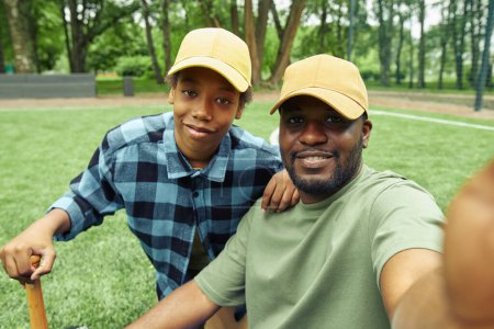 Téléchargez les photos : Portrait selfie de papa et fils heureux en casquettes pendant leur match de baseball dans le parc à l'extérieur - en image libre de droit