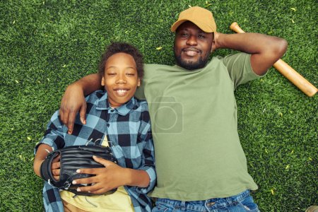 Téléchargez les photos : Vue en angle élevé du père embrassant son fils alors qu'ils gisaient sur l'herbe verte et se reposant après le match de baseball - en image libre de droit