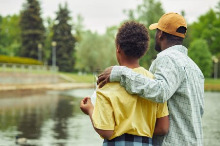 Téléchargez les photos : Vue arrière de papa et fils profitant du beau paysage qui regarde la rivière pendant leur promenade dans le parc - en image libre de droit