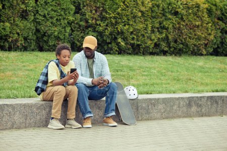 Téléchargez les photos : Garçon montrant des photos sur téléphone portable à son père alors qu'ils se reposent à l'extérieur après le skateboard - en image libre de droit