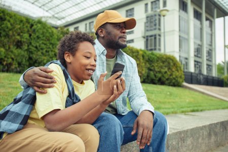 Téléchargez les photos : Père africain profiter du temps avec son fils pendant leur promenade à l'extérieur alors qu'il est assis à jouer au jeu sur téléphone mobile - en image libre de droit