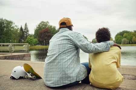 Téléchargez les photos : Vue arrière de papa embrassant son fils et ils se reposent ensemble après le skateboard en regardant la belle vue sur la rivière - en image libre de droit