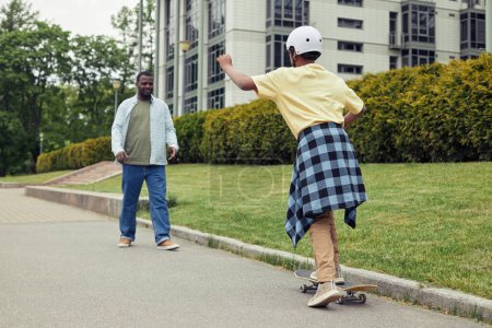 Téléchargez les photos : Africain garçon en casque équitation sur skateboard le long de la route avec son père alors qu'il lui apprend - en image libre de droit