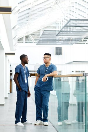 Téléchargez les photos : Portrait complet de deux médecins multiethniques portant l'uniforme debout dans le hall de la clinique moderne et bavardant - en image libre de droit