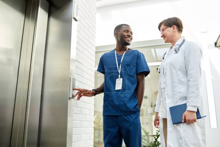 Téléchargez les photos : Portrait en angle bas de deux médecins en attente d'ascenseur dans un bâtiment hospitalier moderne et bavardant joyeusement - en image libre de droit