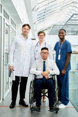 Téléchargez les photos : Portrait complet groupe diversifié de médecins posant avec patient en fauteuil roulant à la clinique moderne, tous regardant la caméra et souriant - en image libre de droit