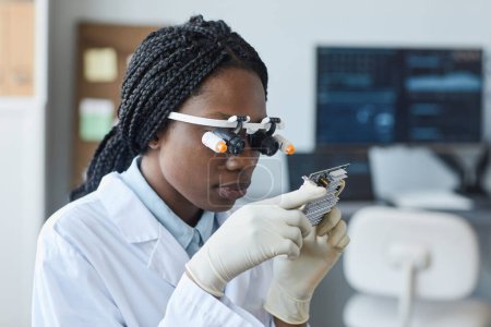 Photo for Portrait of young African American woman wearing magnifying visor and inspecting hardware part in engineering laboratory - Royalty Free Image
