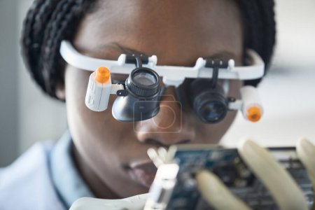 Téléchargez les photos : Gros plan d'une jeune femme noire portant une visière grossissante et inspectant une pièce de quincaillerie dans un laboratoire d'ingénierie - en image libre de droit