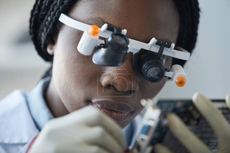 Photo for Closeup of young black woman wearing magnifying glasses and inspecting hardware part in engineering lab - Royalty Free Image