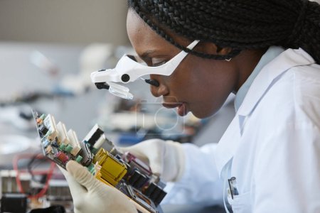 Photo for Side view portrait of black female scientist wearing magnifying glasses and inspecting electronic parts in quality control lab - Royalty Free Image