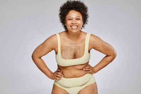 Photo for Minimal waist up portrait of carefree black woman wearing underwear and posing with hands on hips against grey background, copy space - Royalty Free Image