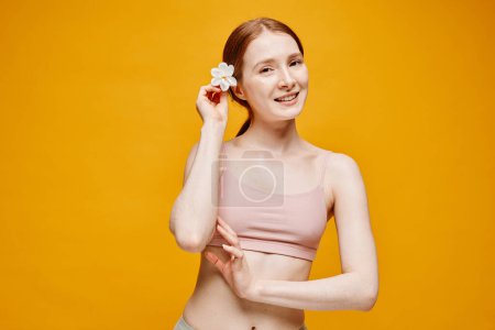 Foto de Cintura mínima hacia arriba retrato de mujer de pelo rojo joven con flor en el pelo de pie sobre el vibrante fondo amarillo de verano, espacio de copia - Imagen libre de derechos