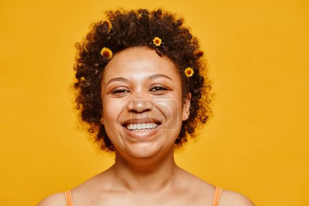 Foto de Primer plano retrato de mujer negra feliz con flores en el pelo sonriendo a la cámara sobre fondo amarillo vibrante - Imagen libre de derechos