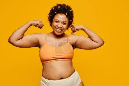 Foto de Retrato de cintura hacia arriba de una poderosa mujer negra sonriendo felizmente mientras posa sobre un vibrante fondo amarillo, positividad corporal - Imagen libre de derechos