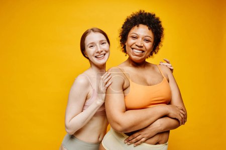 Photo for Waist up portrait of two smiling young women dark skin and fair skin embracing while standing against vibrant yellow background in underwear - Royalty Free Image