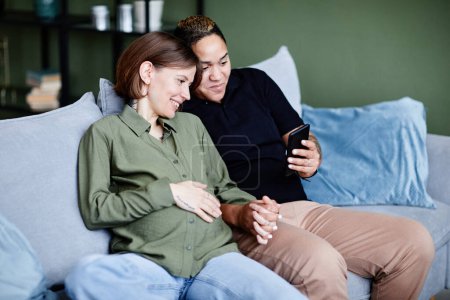Foto de Retrato de pareja gay tatuada con una mujer joven embarazada usando un teléfono inteligente y sonriendo en el interior del hogar, espacio para copiar - Imagen libre de derechos