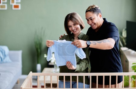 Photo for Waist up portrait of happy lesbian couple expecting baby and preparing crib with baby clothes at home, same sex family, copy space - Royalty Free Image