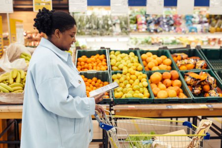 Téléchargez les photos : Vue de côté portrait de femme noire lisant la liste des courses tout en achetant des produits d'épicerie au supermarché, espace de copie - en image libre de droit