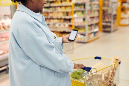 Téléchargez les photos : Gros plan d'une femme noire achetant des produits d'épicerie dans un supermarché et utilisant un smartphone, un espace de copie - en image libre de droit