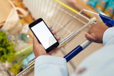 Téléchargez les photos : Gros plan d'une femme noire méconnaissable achetant des produits d'épicerie dans un supermarché et utilisant un smartphone avec un modèle d'écran blanc, un espace de copie - en image libre de droit