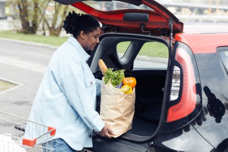 Téléchargez les photos : Vue de côté portrait de femme noire souriante mettant épicerie dans le coffre de la voiture dans le parking, espace de copie - en image libre de droit