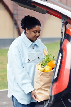Photo for Vertical portrait of young black woman putting grocery bag in car trunk at supermarket parking lot - Royalty Free Image