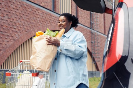 Téléchargez les photos : Portrait à angle bas d'une femme souriante mettant un sac d'épicerie dans un coffre de voiture après avoir fait ses courses au supermarché - en image libre de droit