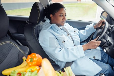 Téléchargez les photos : Portrait d'une femme noire adulte conduisant une voiture avec un sac d'épicerie au premier plan après avoir fait ses courses dans un supermarché - en image libre de droit