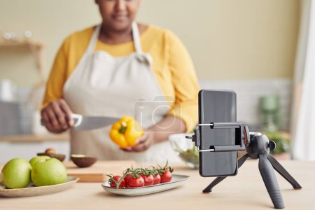 Photo for Black woman filming cooking video in kitchen, focus on smartphone at tripod, copy space - Royalty Free Image