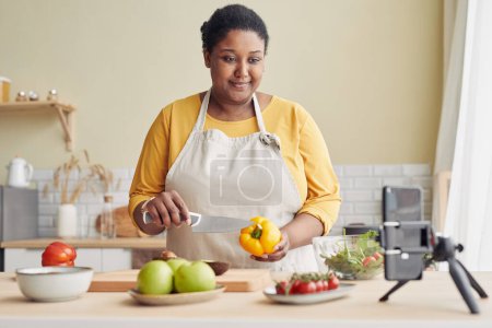 Téléchargez les photos : Portrait taille haute de souriante femme noire cuisine repas sain dans la cuisine et enregistrement vidéo avec smartphone, espace de copie - en image libre de droit