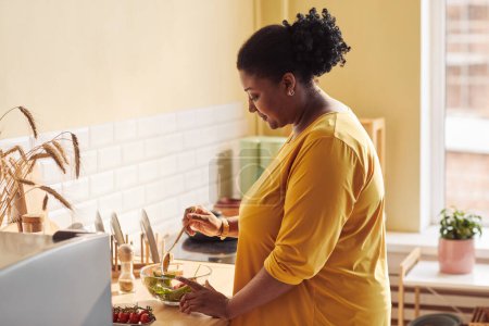 Foto de Retrato de vista lateral de una mujer negra con sobrepeso cocinando comida saludable en la cocina iluminada por la luz del sol, espacio para copiar - Imagen libre de derechos