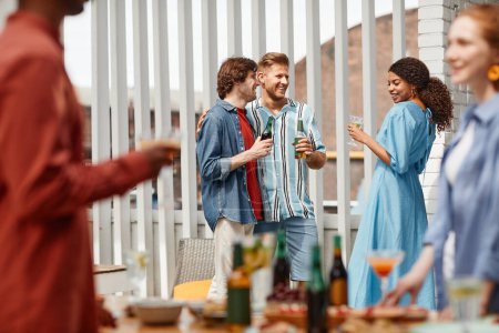 Téléchargez les photos : Groupe diversifié d'amis profitant de la fête en plein air sur la terrasse éclairée par la lumière du soleil - en image libre de droit