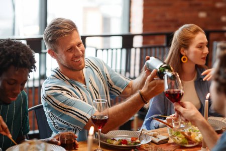 Téléchargez les photos : Portrait d'un homme souriant versant du vin dans un verre tout en profitant d'un dîner entre amis dans un cadre confortable - en image libre de droit