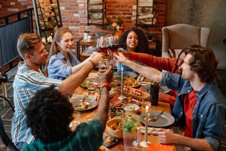Foto de Diverso grupo de amigos tintineando copas de vino mientras celebran juntos durante la cena en un ambiente acogedor - Imagen libre de derechos