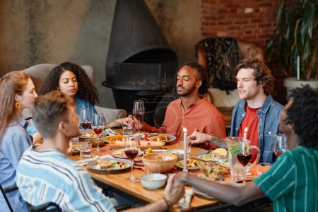 Foto de Diverso grupo de jóvenes tomados de la mano en la mesa y diciendo la gracia durante la cena en un ambiente acogedor - Imagen libre de derechos