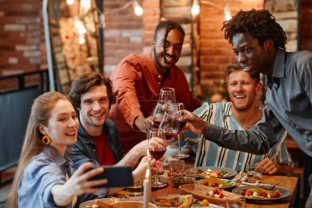 Téléchargez les photos : Divers groupes d'amis prenant des photos selfie à table pendant le dîner et cliquetis de lunettes - en image libre de droit