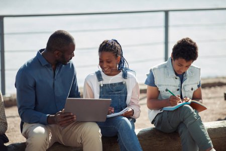 Téléchargez les photos : Groupe d'enfants noirs souriants avec professeur utilisant un ordinateur portable ensemble pendant les cours de plein air à l'école d'été - en image libre de droit