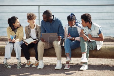 Photo for Full length shot of diverse group of kids with male teacher using laptop outdoors during Summer school lesson - Royalty Free Image