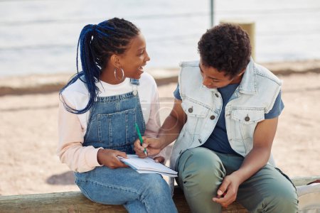 Photo for Portrait of two smiling black teenagers enjoying outdoor class and writing in notebook - Royalty Free Image