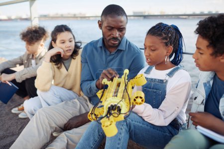 Téléchargez les photos : Portrait de jeune fille noire tenant le modèle de robot pendant la classe d'ingénierie en plein air avec professeur masculin aidant - en image libre de droit